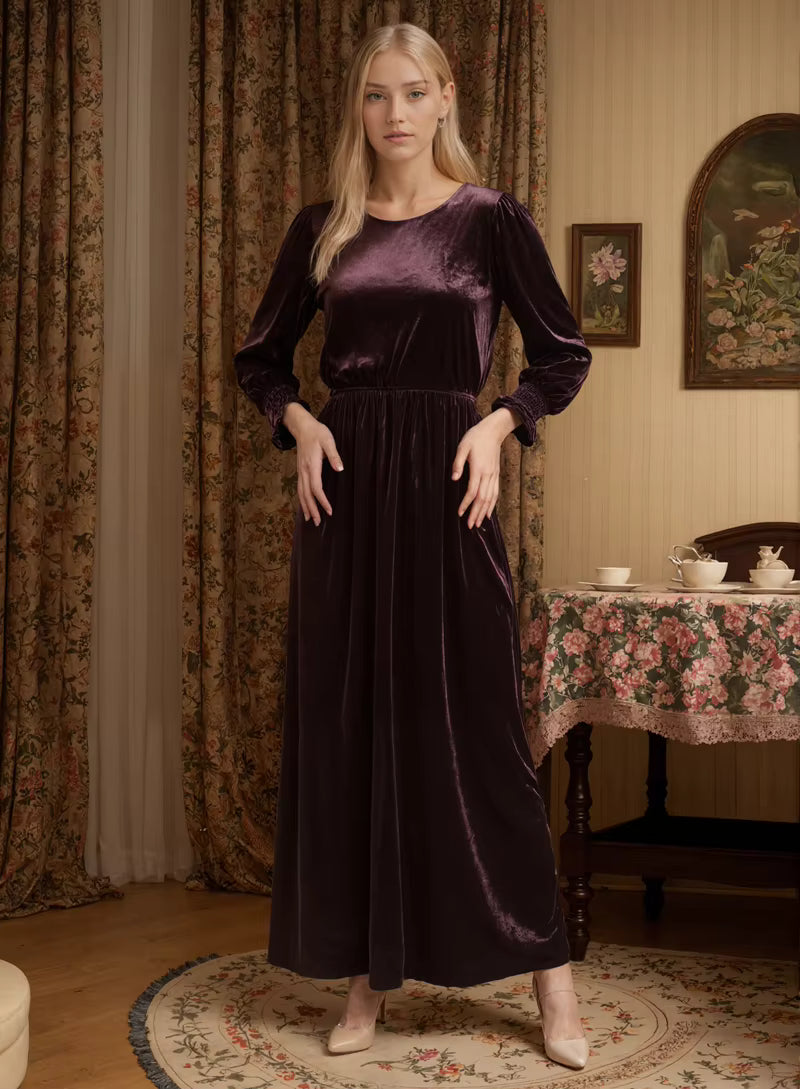 Woman in a long purple velvet dress standing in a room with floral curtains and a table.