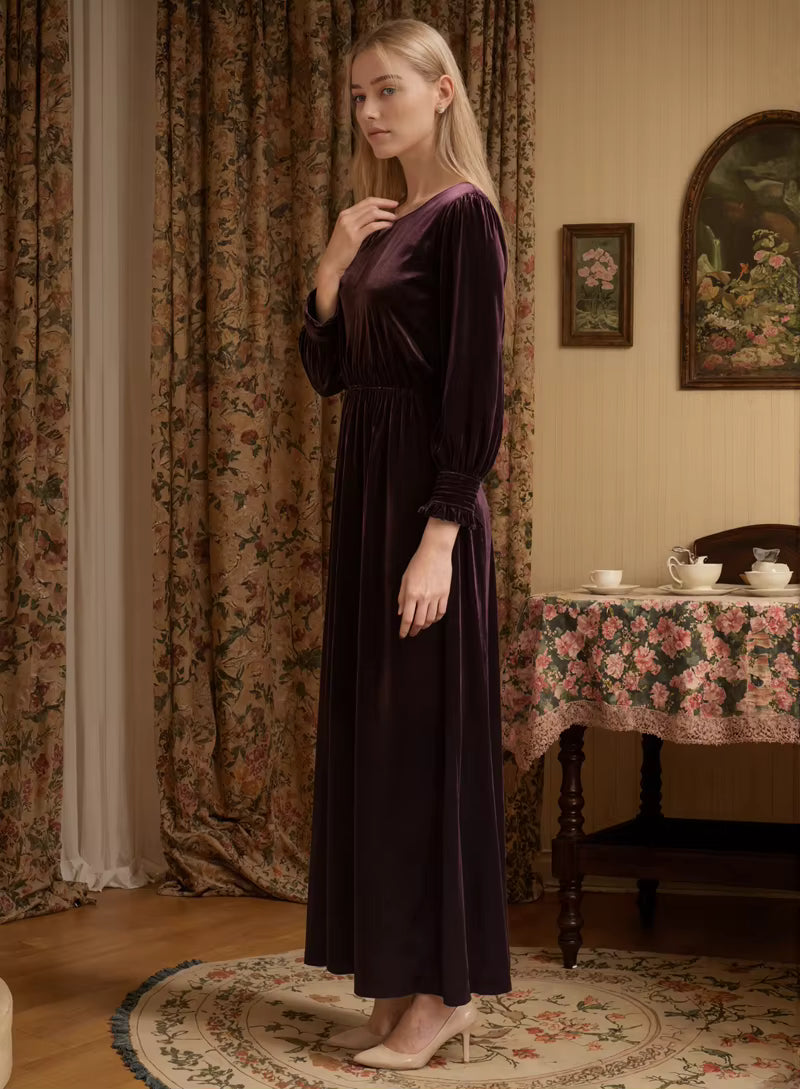 Woman in a dark purple dress standing in a vintage-style room with floral curtains and tablecloth.