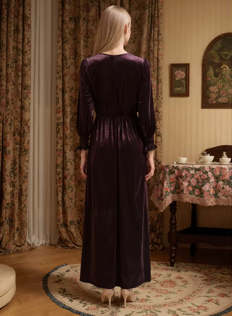 Woman in a long, dark purple dress standing in a room with floral curtains and a table.