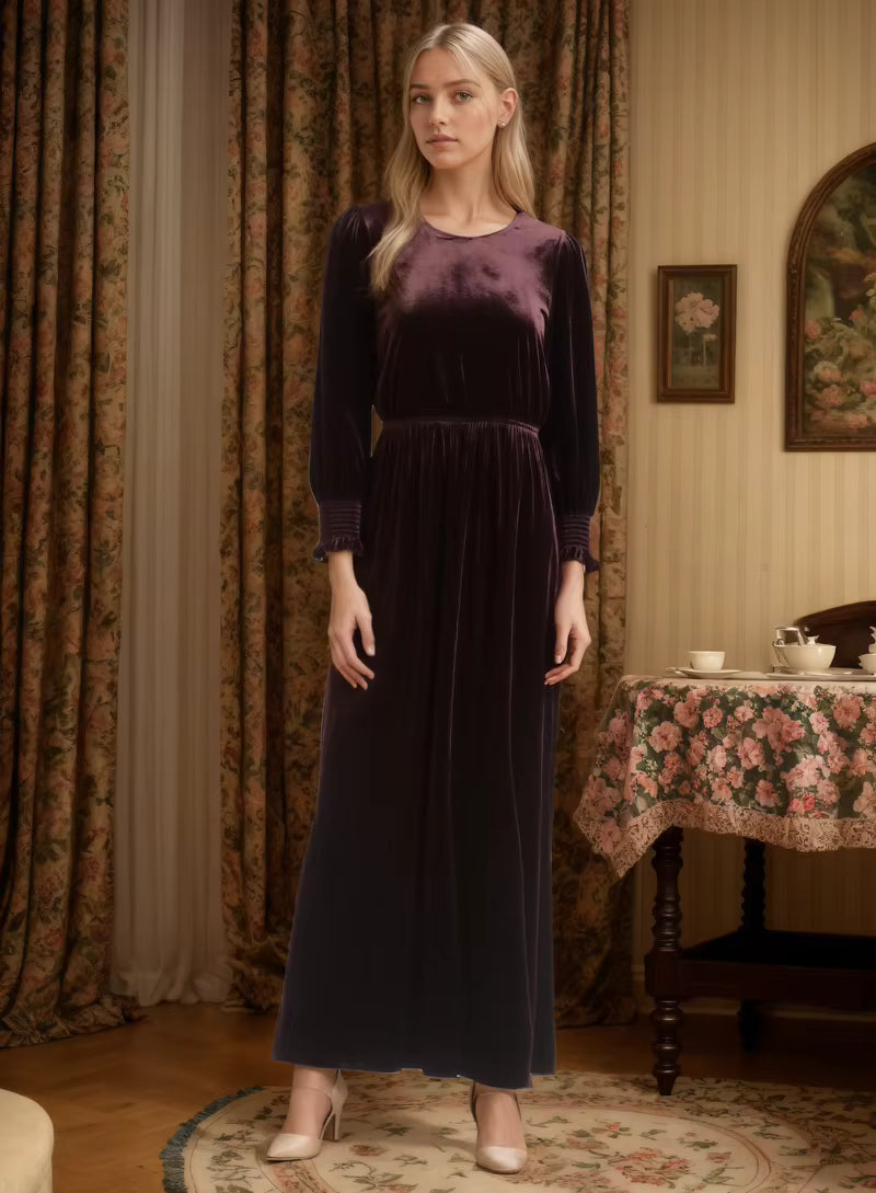 Woman wearing a long dark purple dress in an elegant room with floral curtains and tablecloth.